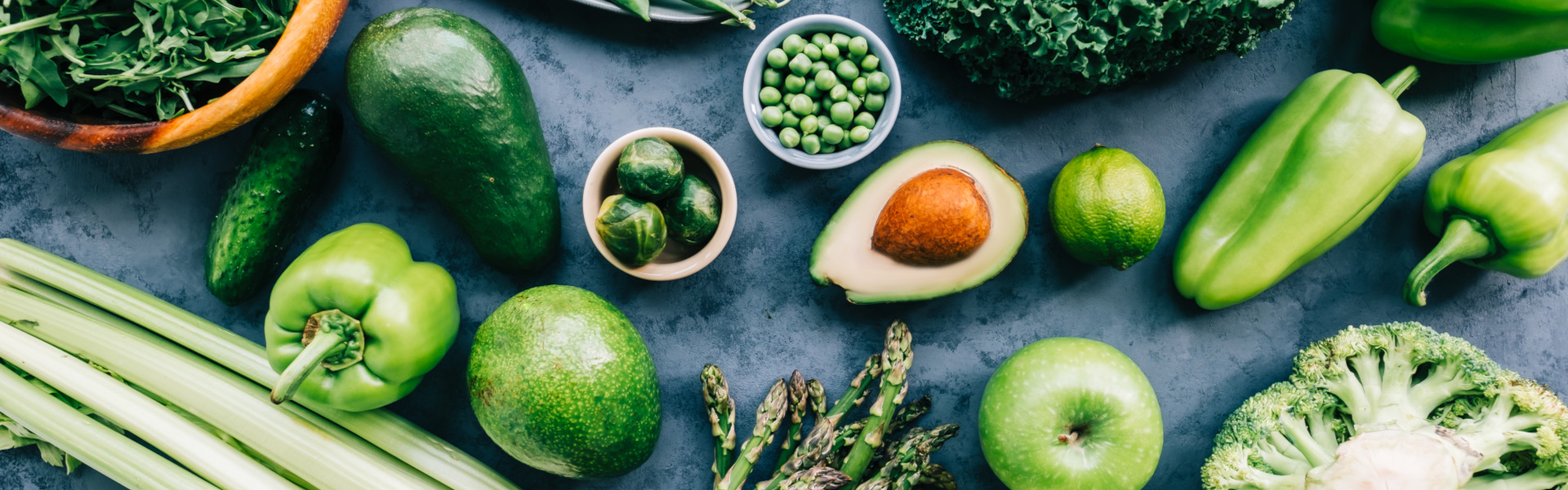 Frisches grünes Obst und Gemüse (Avocado, Erbsen, Rosenkohl, Spargel, Paprika, Apfel) auf dunkler Unterlage.