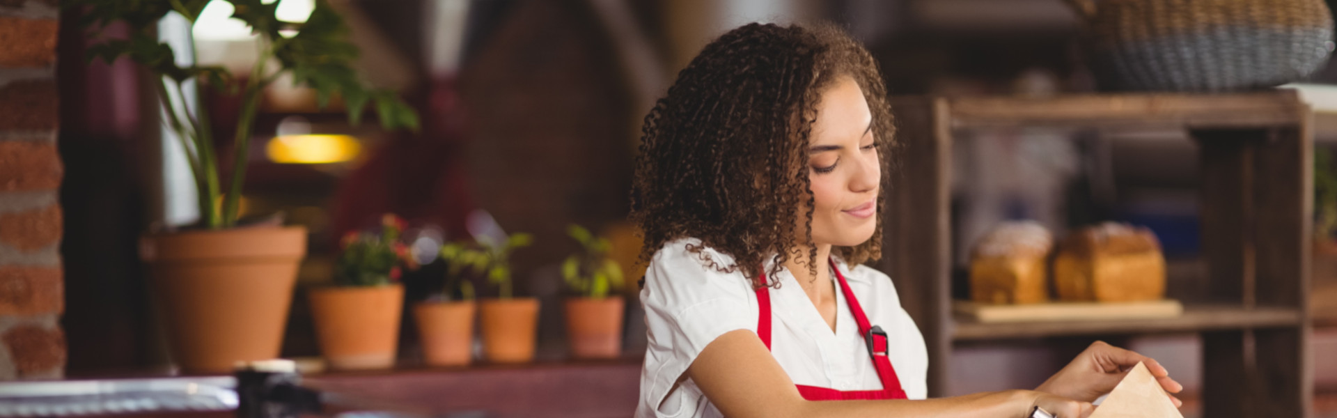 Mitarbeiterin in roter Schürze packt frische Brote aus einem großen Korb in eine Papiertüte in der Bäckerei.